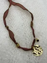 Collar con cinta de seda color marrón, adornado con cuentas doradas y un dije circular dorado con textura irregular.