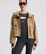 Campera corta color marrón claro, confeccionada en sarga de algodón y poliamida, con cuello alto, capucha y bolsillos delanteros con solapa. Tiene cierre frontal con botones a presión y mangas largas con puños elásticos.