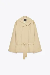 Trench color beige con cuello de solapa subido y manga larga. Tiene bolsillos laterales, forro interior y cinturón de lazo en el mismo tejido. El cuello se cierra con un botón.