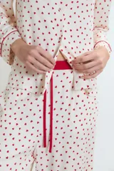 Pijama de dos piezas, compuesto por una camisa de manga larga y pantalón largo, ambos en tejido de viscosa color blanco con estampado integral de corazones rojos. La camisa tiene ribetes rojos en cuello, puños y abertura frontal. El pantalón tiene un fajín ancho color rojo liso.