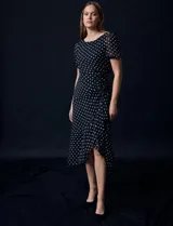 Vestido midi de gasa color negro con lunares blancos, con mangas cortas, escote redondo y ruedo asimétrico. Presenta un frunce lateral con lazo de ajuste.