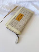 Billetera grande con cierre metálico, color beige y crema. El frente presenta un tejido texturado con patrón de rayas horizontales en tonos claros, ribetes en charol color crema y un parche de cuero marrón con la marca grabada.