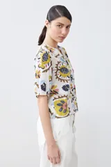 Camisa blanca con estampado floral en tonos azul, amarillo, rosa y marrón. Tiene cuello mao, mangas cortas abullonadas y cierre frontal con botones.