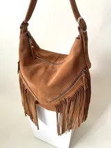 Bolso tipo hobo color tostado, hecho de cuero con flecos largos en el frente y dos bolsillos con cierre. Tiene una manija regulable con hebillas metálicas.