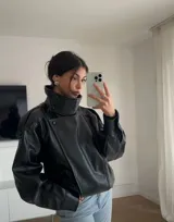 Chaqueta negra de efecto cuero, estilo bomber o motociclista, con cuello alto abotonado y cierre frontal asimétrico. Presenta detalles de costuras verticales y hombreras pequeñas. El bajo es elástico.