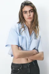 Camisa celeste de popelín con cuello solapa, escote pico y manga corta. Presenta dos bolsillos delanteros de parche y cierre frontal con botones.