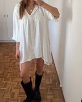 Vestido camisero blanco, holgado, con cuello y botones frontales. Presenta frunces bajo el canesú y mangas tres cuartos con elástico en los puños. El ruedo es asimétrico, más corto adelante que atrás.