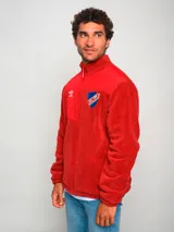 Campera polar roja con cierre frontal, cuello alto y bolsillos laterales. Presenta un panel liso en la parte superior del pecho y un bolsillo con cierre en el lado izquierdo. Incluye el logo de Umbro bordado en el pecho izquierdo y el escudo del Club Nacional de Football bordado en el pecho derecho.