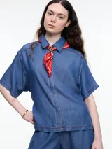 Camisa de manga corta confeccionada en denim liviano de tencel, color azul, con tacto ultra suave y gran fluidez. Su calce es cuadrado y relajado. Presenta cierre frontal con tapeta escondida y costuras en color contraste que resaltan las líneas de construcción.