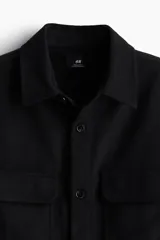 Sobrecamisa negra de corte recto, con cuello clásico, cierre frontal con botones, dos bolsillos de parche con solapa en el pecho, mangas largas con puños abotonados y bajo redondeado.