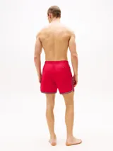 Short de baño rojo con cintura elástica ajustable con cordón blanco. Tiene dos bolsillos laterales y un bolsillo trasero de parche. El bajo presenta una franja con los colores azul marino, blanco y rojo.