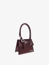 Bolso estructurado de piel lisa color borgoña, con asa reforzada de hebilla metálica, solapa magnética y asa bandolera ajustable y extraíble. Cierre de mosquetón grabado, bolsillo interior y logotipo frontal. Forrado completamente en algodón.