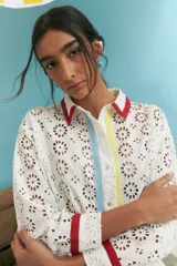 Camisa blanca oversize de algodón con bordado calado tipo broderie, cuello camisero con ribete rojo, cierre frontal con botones y mangas largas con puños con ribete rojo. Presenta una franja vertical con ribete celeste y otra con ribete amarillo.