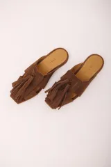 Zueco de cuero gamuzado color marrón, con punta cuadrada y detalle de gran borla de flecos en el empeine. Presenta aberturas laterales y suela plana.