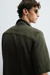 Camisa verde militar de corte relaxed fit, con cuello bowling, manga corta, bajo con aberturas laterales y cierre frontal de botonadura.