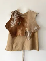 Chaleco pechera de cuero color marrón con manchas blancas, de corte irregular y diseño rústico.