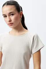 Remera color beige de punto acanalado con silueta relajada y manga corta raglán. El detalle de ruedo con terminación ondulada le aporta un aire fresco y juvenil.