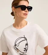 Remera blanca de algodón con cuello redondo y bordado de una cara de mujer con caravana dorada.