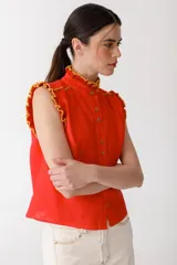 Blusa color mostaza sin mangas, con cuello alto y detalle de volados rojos en cuello, sisas y ruedo. Cierre frontal con botones.