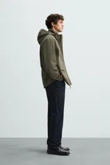 Parka verde militar de tejido técnico ligero resistente al viento, con cuello alto, capucha y manga larga con puños ajustables. Tiene bolsillos de plastrón con solapa en la cadera, bolsillo interior y bajo ajustable con elásticos. El cierre frontal es con cremallera oculta por solapa con botones a presión.