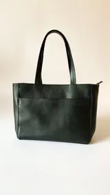 Cartera tipo tote de cuero graneado negro, con textura uniforme y suave al tacto. Tiene manijas de 55 cm, bolsillo frontal con broche de imán, bolsillo trasero con cierre metálico, cierre metálico principal, forro interior, tres bolsillos internos (uno con cierre, uno plaqué y uno tipo sobre con solapa y botón).
