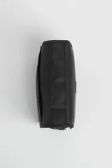 Necesser de viaje negro con cierre de cremallera y asa de transporte. Tiene un parche rectangular con el logo de un avión y la palabra "TRAVEL".