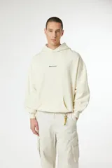 Hoodie color crema o blanco roto, de corte holgado y manga larga, con bolsillo canguro frontal y un pequeño logo bordado en el pecho.