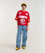 Remera roja de algodón con estampado del logo de los Kansas City Chiefs, el nombre del equipo y el número 60 en blanco. Tiene mangas cortas con rayas amarillas y rojas.