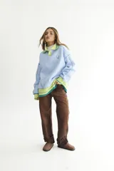 Buzo celeste con cuello polo y puños en rib color verde lima. Tiene un parche con el logo de la marca en el pecho y rayas horizontales en el ruedo.