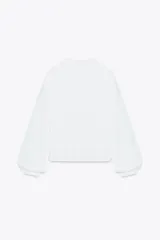 Camisa blanca de cuello redondo y escote pico, con mangas largas abullonadas y detalle de puntillas combinadas a tono en el delantero. Cierre frontal con botones forrados.
