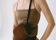 Bolso pequeño de piel bovino color marrón oscuro, con acabado en serraje (gamuza). Presenta un forro interior de algodón y cierre principal de cremallera. Incluye un asa larga ajustable.