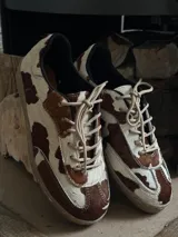Sneakers de cuero con estampado de vaca en tonos marrón y blanco, con cordones beige y suela de goma color caramelo.