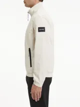 Campera Calvin Klein color beige claro, confeccionada en nailon arrugado reciclado. Tiene cuello alto, puños y bajo elásticos, cierre con cremallera, bolsillos con cremallera en la cintura, abertura de ventilación en la espalda, forro de poliéster reciclado e insignia con el logo de goma de Calvin Klein en la manga.