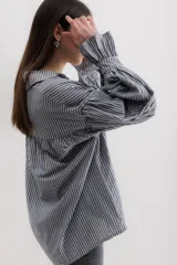 Camisa gris a rayas verticales blancas, de corte amplio, cuello alto con lazo y mangas largas abullonadas con puños elásticos.