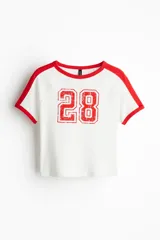 Remera corta blanca de punto acanalado fino de algodón con mangas raglán y ribetes en rojo. Estampado frontal con el número 28 en rojo.