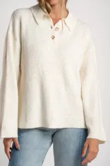 Sweater de tejido de punto color blanco roto o crema, con textura jaspeada. Presenta cuello polo y un escote con tres botones nacarados. El calce es holgado (oversize) y tiene mangas largas.