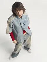 Camisa de manga corta Adidas Premium Essentials, confeccionada en denim azul claro 100% algodón. Diseño inspirado en la ropa de trabajo, con cuello clásico, cierre frontal con botones, bolsillo de parche en el pecho y logo bordado en el mismo tono. Presenta las tres tiras características de la marca en color beige en los hombros.