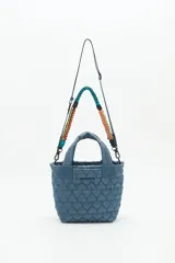 Cartera tote azul de nylon matelassé con diseño de corazones. Tiene manijas cortas y dos correas desmontables, una de cuero sintético y otra de tela trenzada multicolor. Cuenta con cierre superior y dos bolsillos internos.
