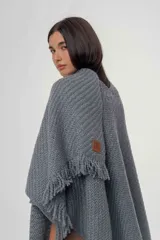 Ruana de punto color gris con diseño de trama diagonal y terminación de flecos en el ruedo. Presenta un pequeño parche decorativo en la parte frontal inferior.