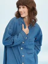 Camisa de denim negro con corte oversize, cuello clásico, cierre frontal con botones metálicos y bolsillo de parche en el pecho.