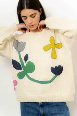 Sweater de lana color crudo con cuello redondo y mangas largas. Presenta apliques de tela cosidos con forma de flores y figuras geométricas en tonos gris, azul, verde, amarillo y rosa.