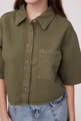 Camisa color verde oliva oscuro, de corte cropped y holgado, con cuello clásico, manga corta y bolsillo en el pecho.