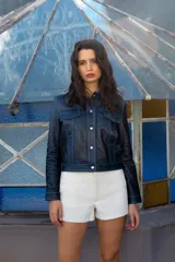Chaqueta corta de cuero negro con pespuntes azules, cuello clásico, cierre frontal con botones metálicos y bolsillos de parche en el pecho.