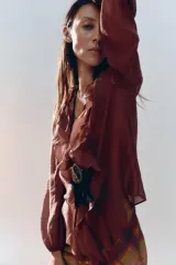 Camisa color crudo confeccionada en tejido con lyocell. Presenta escote en pico, mangas largas abullonadas con elástico en los puños, volantes frontales y cierre frontal con botones.
