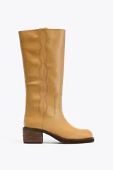 Bota alta de cuero color camel, con caña recta y punta redondeada. Presenta pespuntes decorativos verticales en la parte delantera y un tacón ancho de madera.