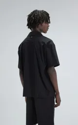 Camisa negra de manga corta estilo técnica, confeccionada en microfibra liviana. Tiene doble bolsillo frontal con cierre de cremallera y broches en lugar de botones.