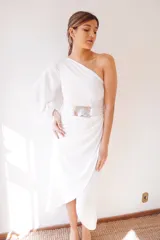 Vestido largo blanco de seda, con un hombro descubierto, manga larga abullonada y falda irregular con drapeado lateral. Incluye cinturón blanco con hebilla de alpaca y nácar.