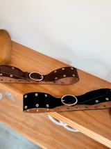 Cinturón ancho de doble capa, con capa superior de gamuza negra o marrón y capa inferior de cuero color camel. Presenta múltiples ojales metálicos y hebilla ovalada metálica de color dorado.