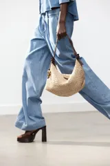 Bolso tipo shopper color beige, con cuerpo trenzado de rafia y asa de hombro de cuero marrón. Tiene un charm colgante en un lateral y cierre con cremallera.