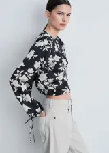 Blusa de satén color negro con estampado de flores blancas, cuello redondo, cierre frontal con botones, manga larga con puños elásticos y fruncidos, y corte recto.